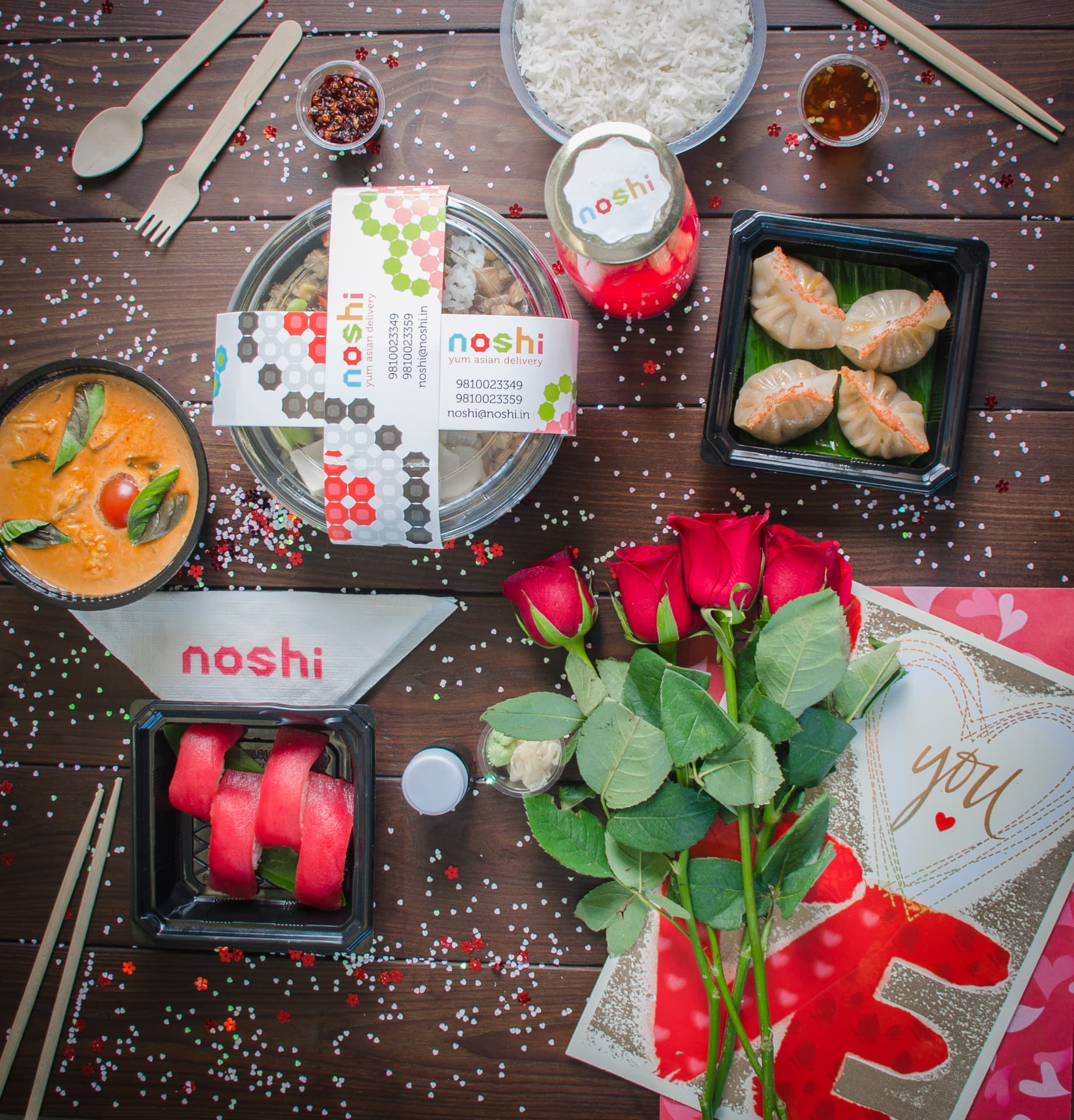 Flat-lay of an Asian delivery spread with curry, dumplings, sushi and roses on a wooden table