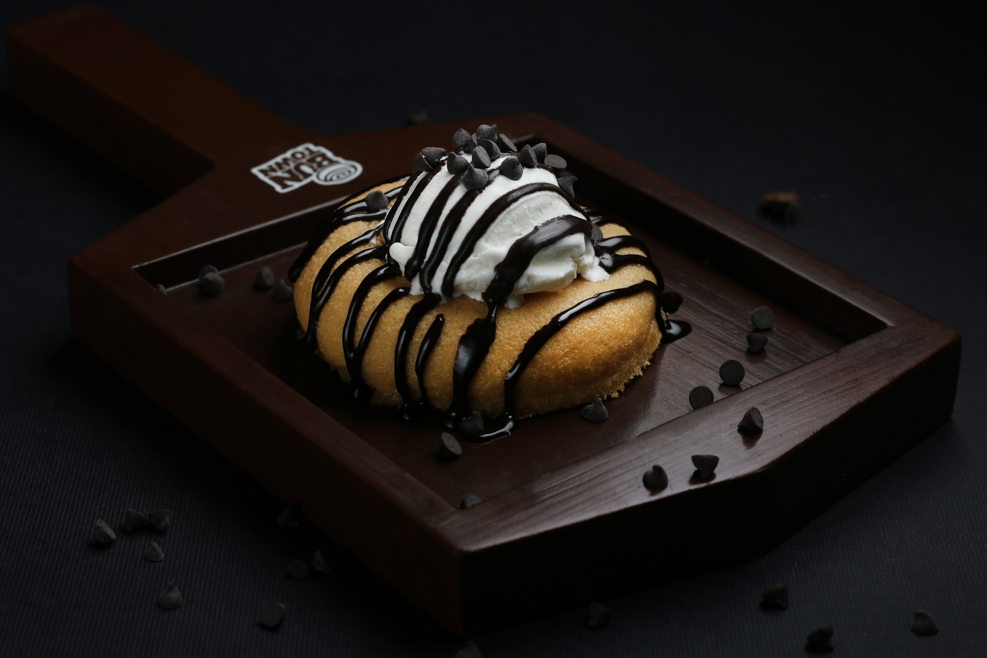 Dessert bun topped with ice cream and chocolate drizzle on a dark wooden board